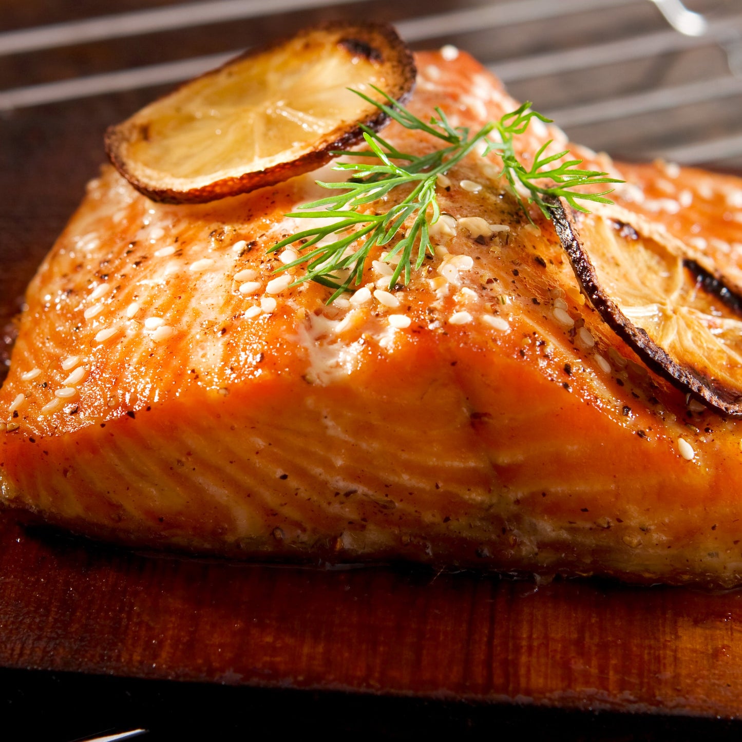 A cooked salmon fillet seasoned with roasted sesame seeds and fresh green herbs is served on a wooden board with grilled lemon slices.