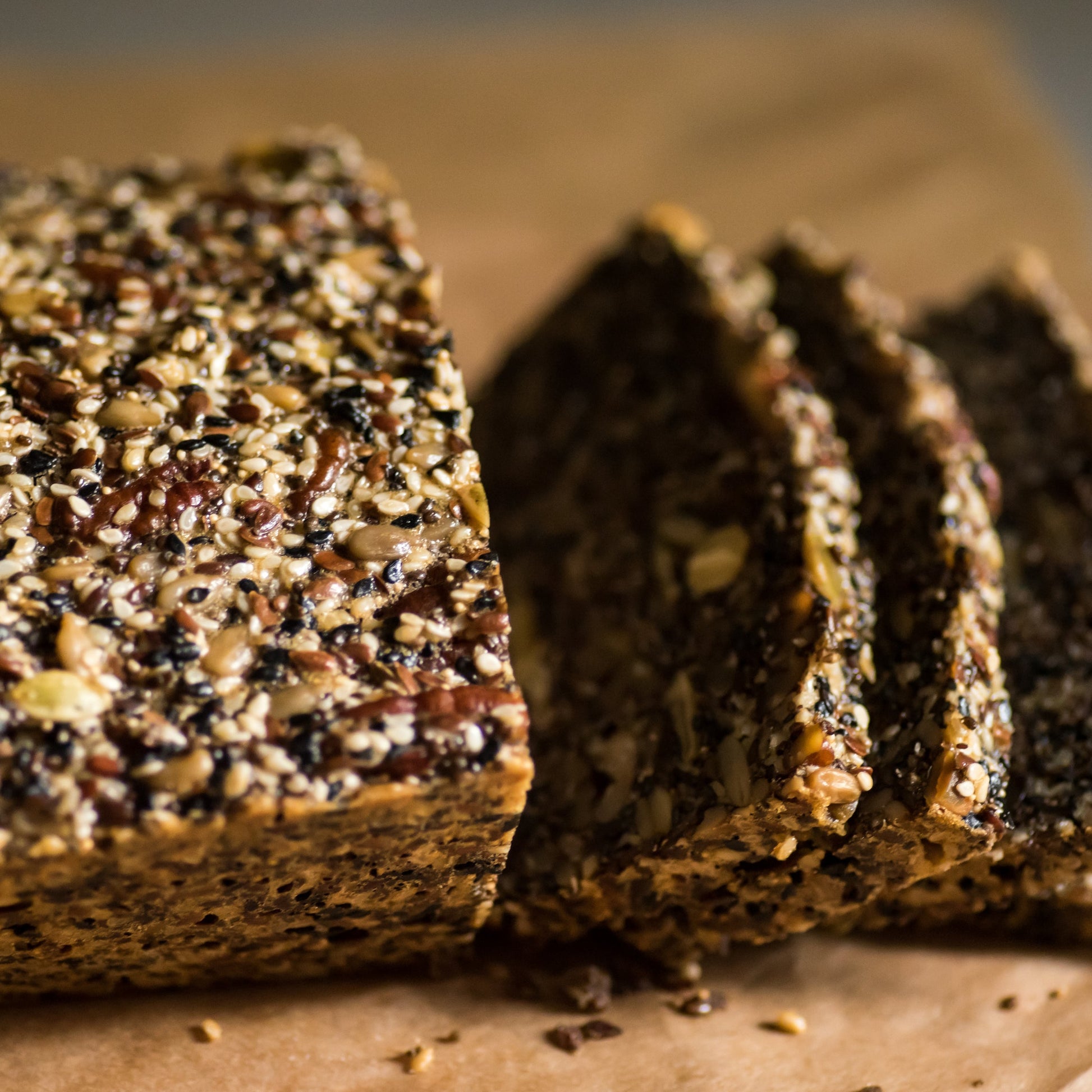 Slices of rustic seeded bread sit on a wooden surface, with sunflower seeds and other seeds visible throughout the crust and crumb.