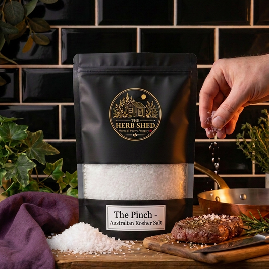 A black stand-up pouch of The Herb Shed The Pinch Australian Kosher Salt sits on a wooden surface with loose white kosher salt and grilled meat, while a hand sprinkles salt from above, surrounded by green foliage, copper cookware and purple fabric against a black tiled kitchen background.