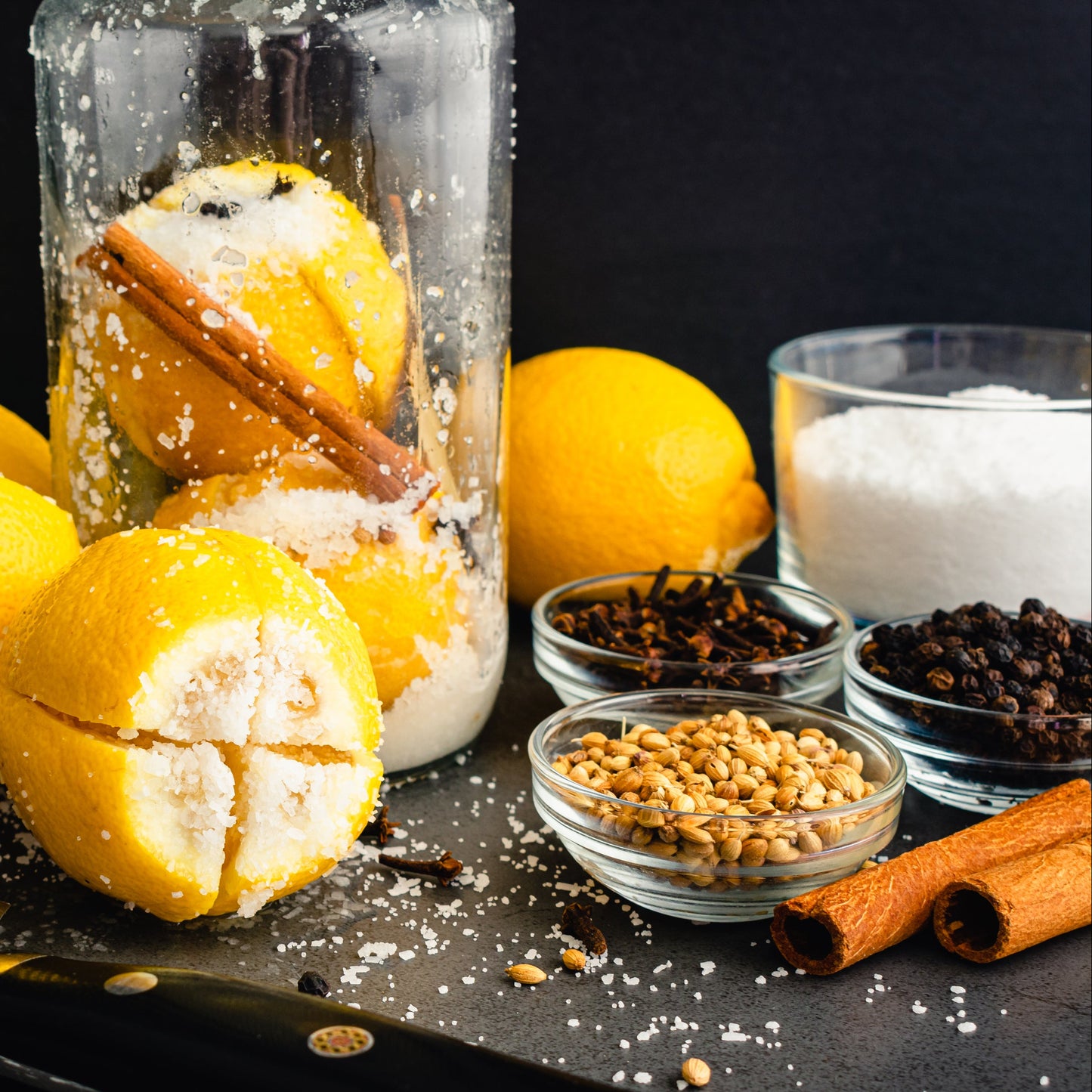 Fresh lemons, Australian kosher salt and whole spices are arranged beside a glass preserving jar, showing the ingredients used to make preserved lemons.