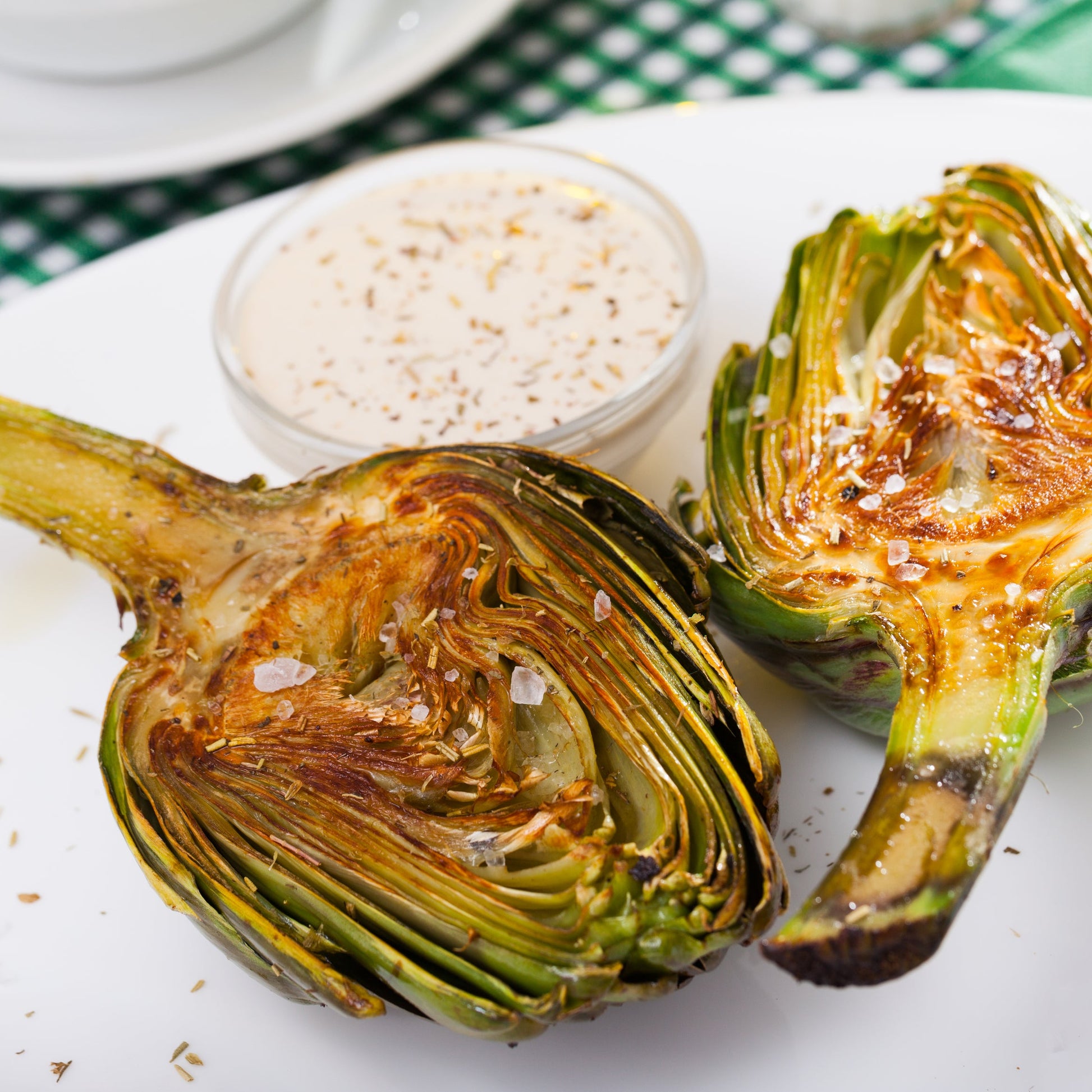 Roasted halved artichokes seasoned with Australian kosher salt are served on a white plate with a savoury dipping sauce.