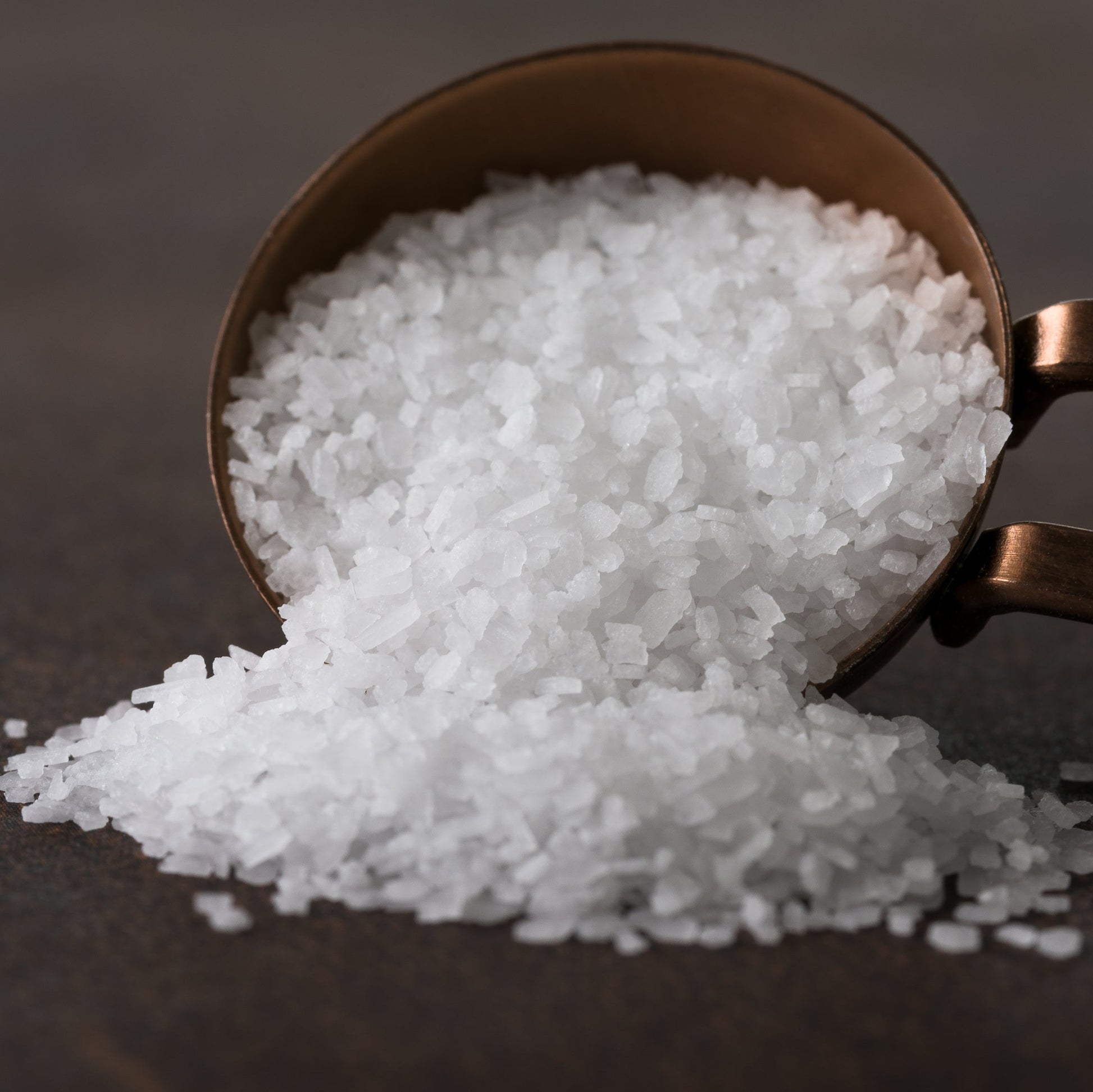 A close-up of coarse Australian kosher salt spilling from a teaspoon onto a dark surface, showing the large white crystals and coarse texture of the salt.