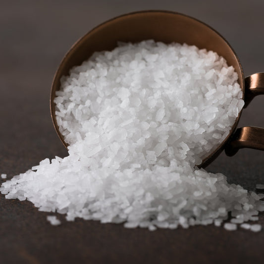 A close-up of coarse Australian kosher salt spilling from a teaspoon onto a dark surface, showing the large white crystals and coarse texture of the salt.
