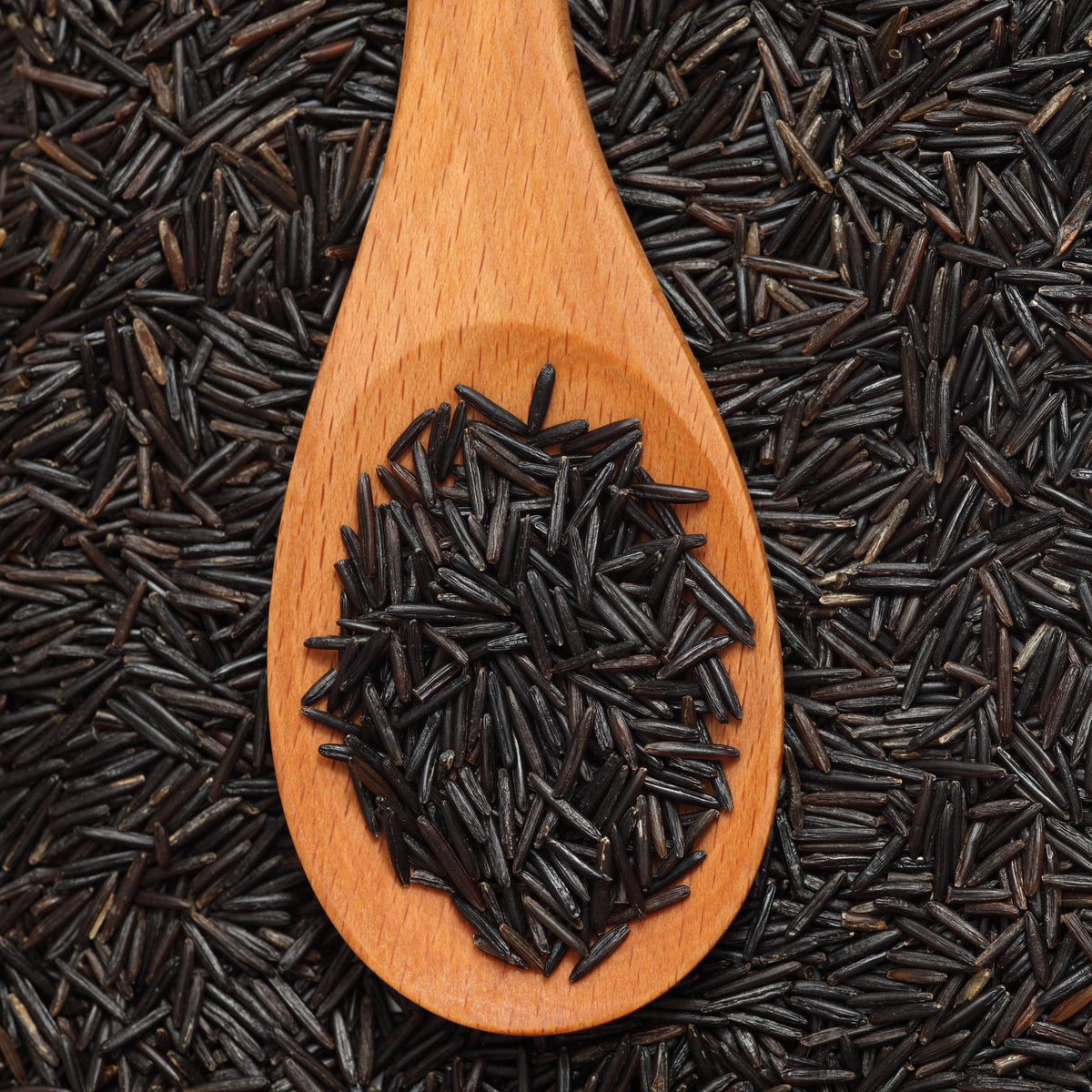 Uncooked wild rice fills a wooden spoon surrounded by loose grains, showing the long, narrow shape and dark colouring of the grain.
