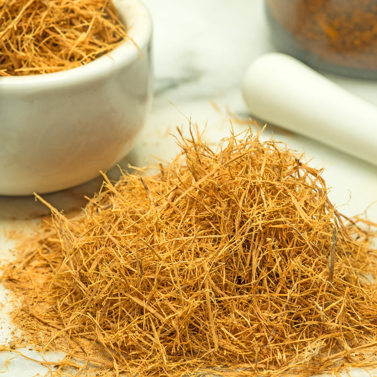 A pile of Wildcrafted Cats Claw shredded bark sits beside a small bowl and mortar and pestle, showing the fine dried strands and their natural golden-brown colouring.