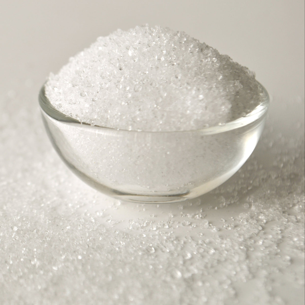A clear glass bowl filled with white xylitol crystals sits on loose xylitol, showing the fine sugar-like texture of the natural sweetener.