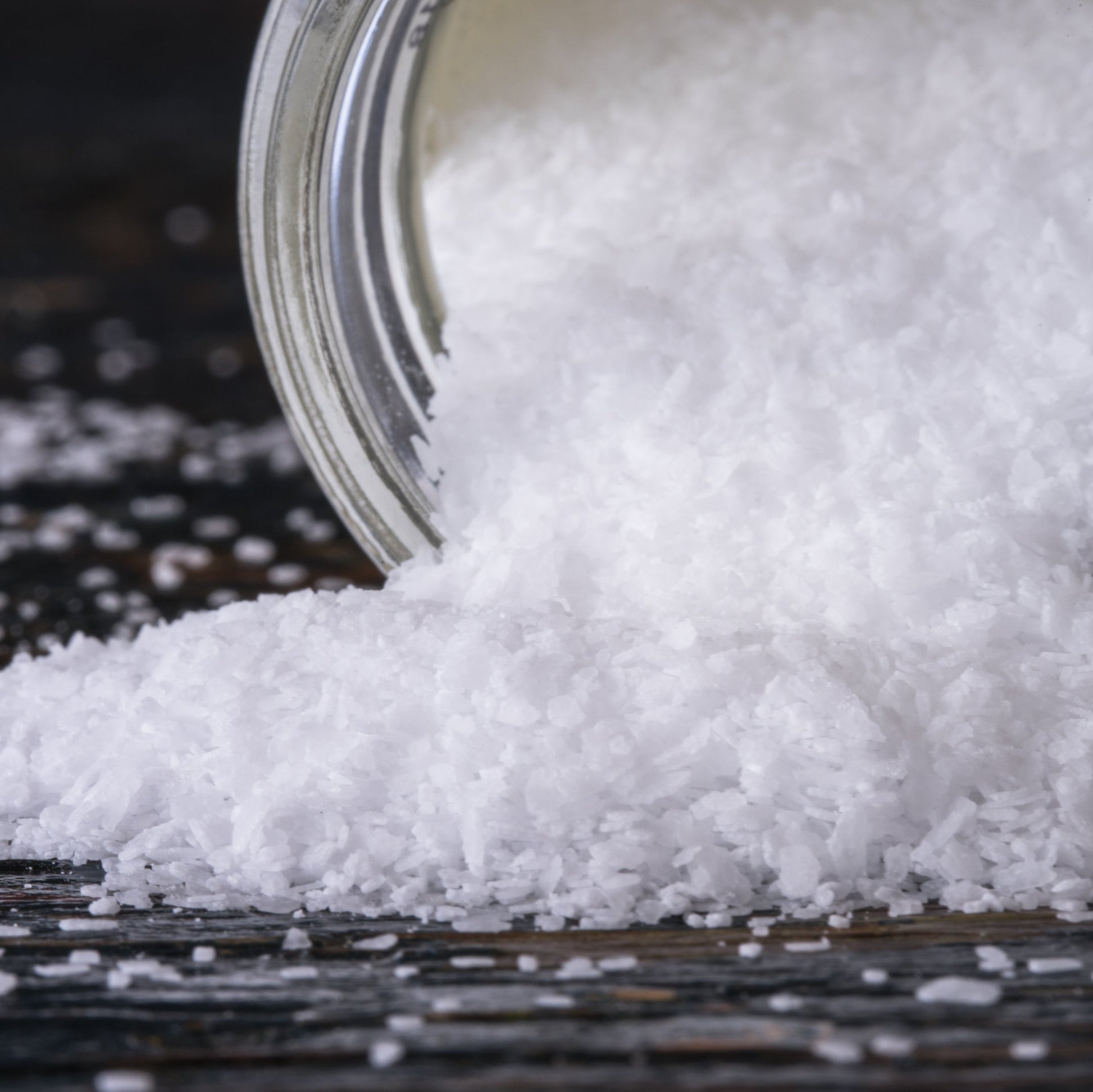White coarse salt crystals spilling from a clear glass jar onto a dark wooden surface.