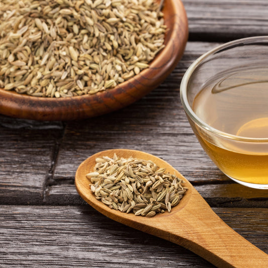Fennel seeds in a wooden spoon beside a glass cup of fennel tea on a rustic wooden table.