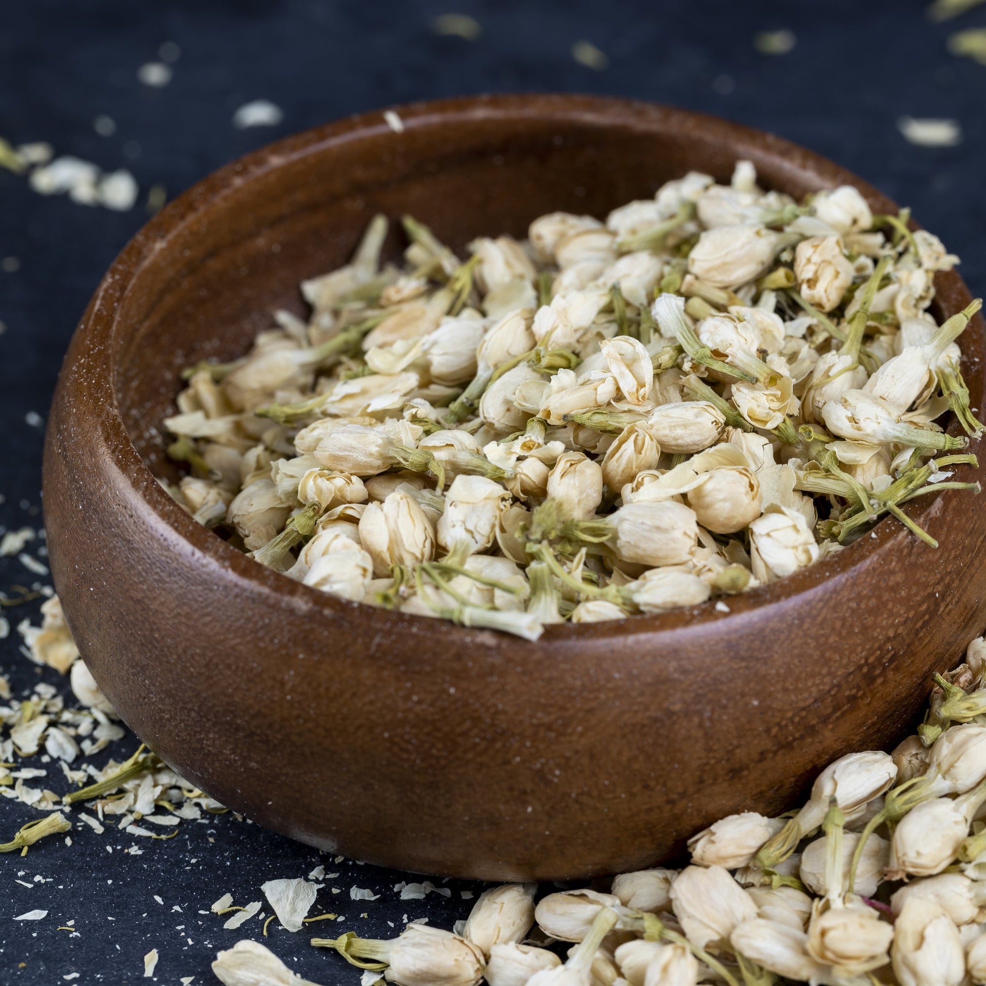 Jasmine flower buds in a brown wooden bowl on a dark blue table, with buds scattered and spilling out – delicate fragrant blossoms from The Herb Shed Wollongong