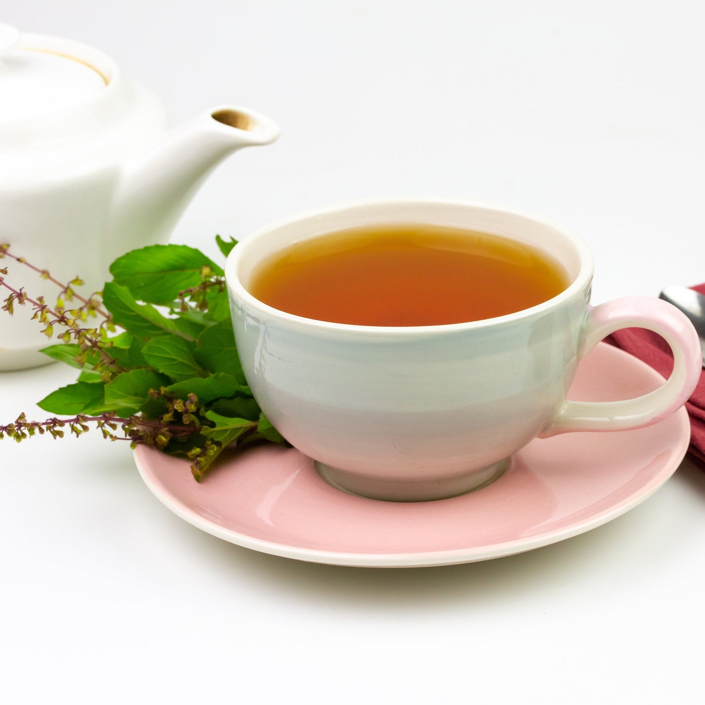 Cup of tulsi tea on a pink saucer with fresh tulsi leaves and a white teapot on a white background – calming herbal tea from The Herb Shed Wollongong